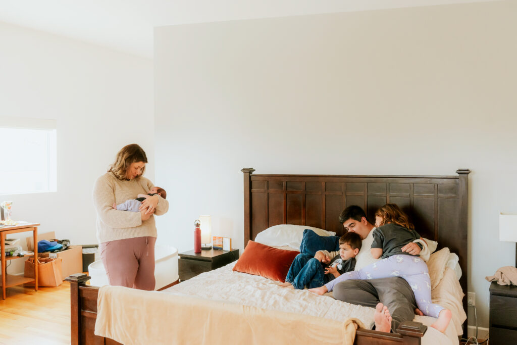 family gathered on the bed while mom stands holding newborn baby